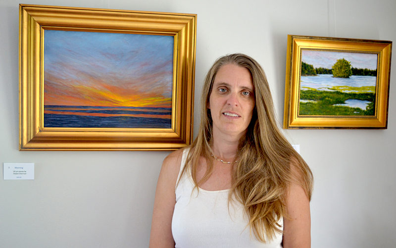 Pemaquid Watershed Association Executive Director Jennifer Hicks is flanked by two of Helen Warner's oil paintings that are part of Warner's show, "Our Natural World," currently hanging on the walls of the PWA's new office at 584 Main St., Damariscotta. At left is "Morning"; on the right is "Looking Upstream." (Christine LaPado-Breglia photo)