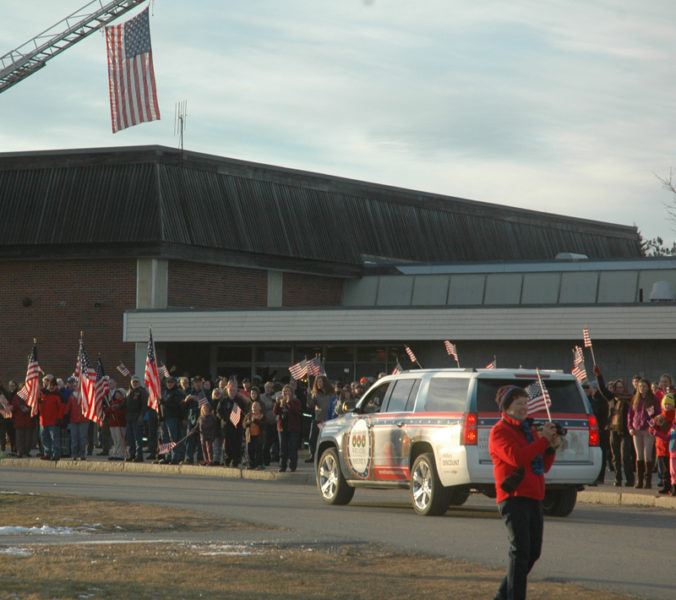 A crowd of people waving flags welcomes the Wreaths Across America convoy to Medomak Valley High School. (Alexander Violo photo)