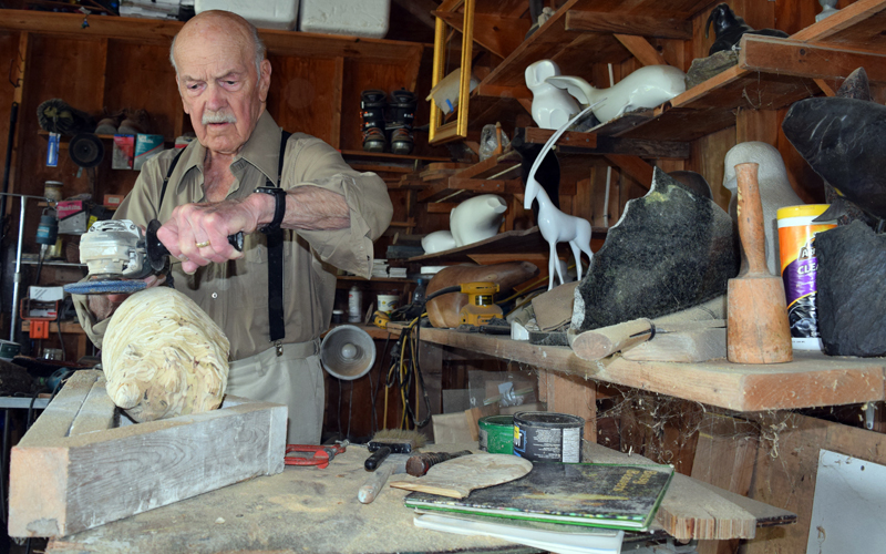 Ralph Moxcey uses an electric grinder to shape a wooden beaver sculpture in his Bremen garage. Some of his previous sculptures are on the shelves beside him. (J.W. Oliver photo)