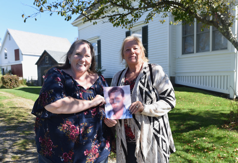 Sisters Carolyn Mattern (left) and Liz Michaud hold a photo of their late brother, Sheldon "Skip" Cushing, in front of what was once their great-grandparents' home in Round Pond. (Jessica Picard photo)