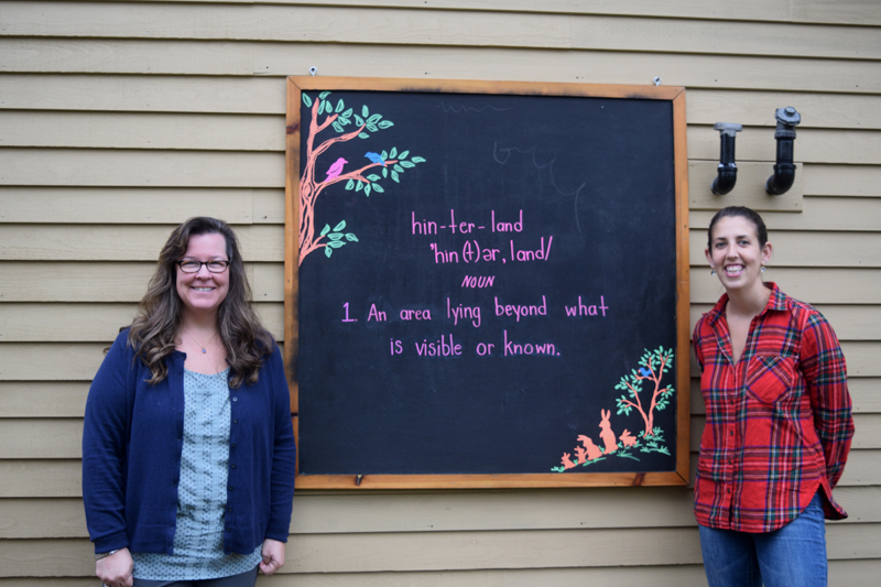 Melissa Vallieres (left) and Brandi Grady stand in front of a sign outside their Sheepscot Valley Hinterland Preschool. The sign defines hinterland as "an area lying beyond what is visible or known." (Jessica Clifford photo)