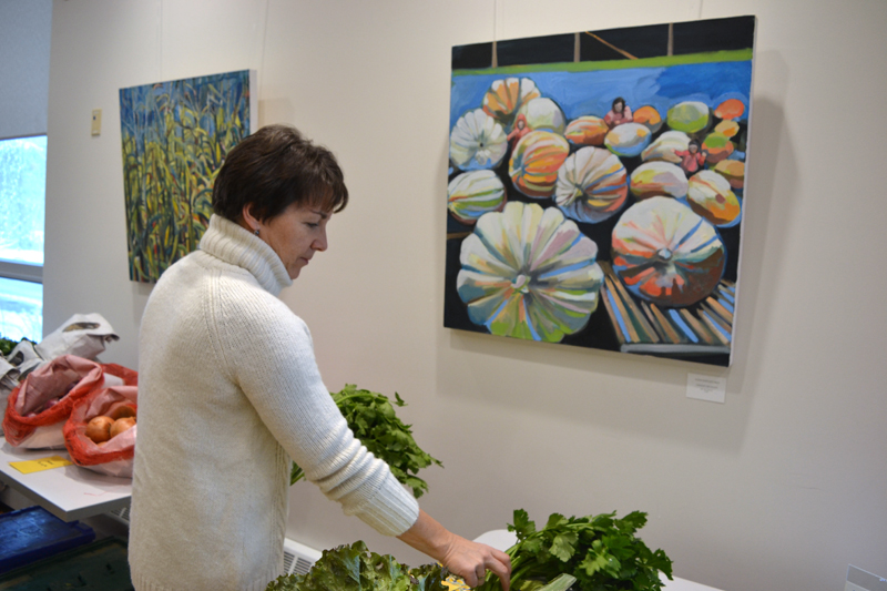 Susan Bartlett Rice's paintings "Maize" (left) and "Amongst the Giants" serve as a backdrop as Central Lincoln County YMCA Membership and Marketing Director Leslie Gomes chooses a bunch of celery from the Morning Dew Farm table at the Y on Friday, Nov. 16. (Christine LaPado-Breglia photo)