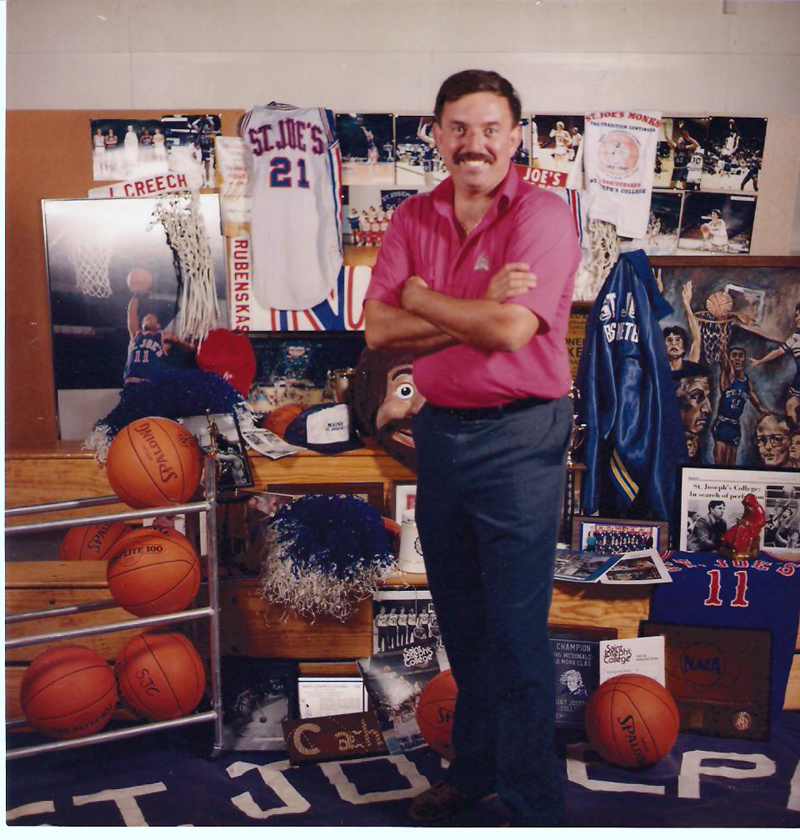 Rick Simonds poses for a photo in front of a collection of St. Joseph's College men's basketball memorabilia. The photo was the cover of the school's 1989-1990 media guide. (Photo courtesy St. Joseph's College Athletics)