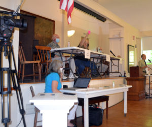 Westport Island Third Selectman Ross Norton raises his card in opposition to accepting a section of North End Road as a town way during annual town meeting at the historic town hall, Saturday, July 18. COVID-19 precautions at the meeting included face masks, Plexiglas barriers in front of the moderator and selectmen, and audiovisual equipment to connect voters at the town hall with those present in the community church next door. (Charlotte Boynton photo)