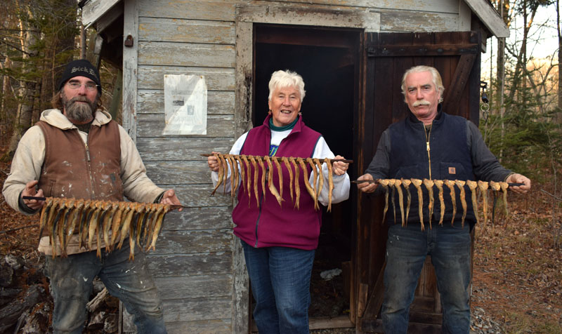 From left: Todd Lincoln, Donna Plummer, and Ken Lincoln hold rods of smoked herring in front of the old smokehouse next to the S Road School in South Bristol. A rod is available for a $5 donation to the South Bristol Historical Society. (Evan Houk photo)