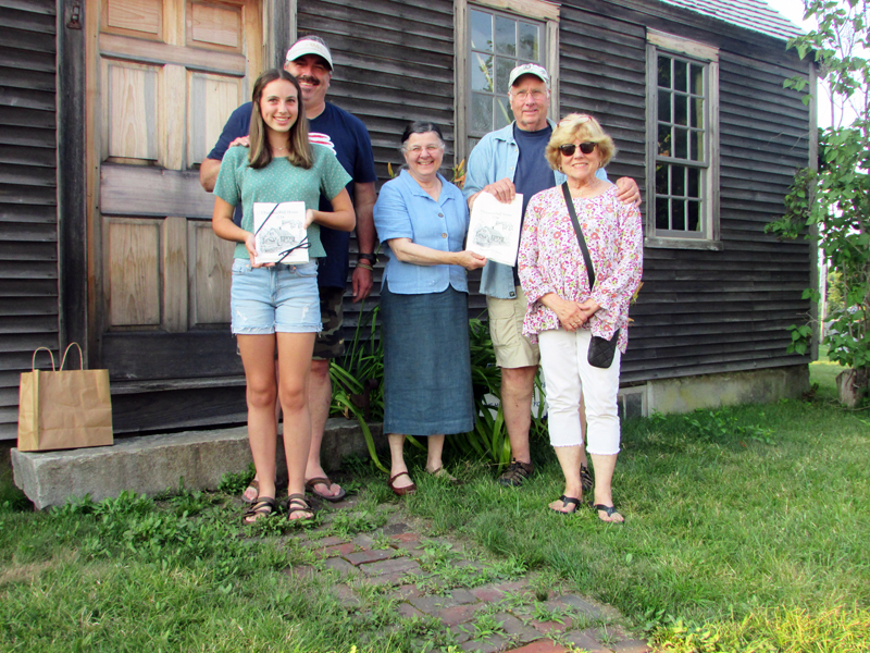 Louise Miller, of the Chapman-Hall House Stewardship Committee, presents Sandi and Dan Day, their son Justin, and his daughter Zofie, with towels printed with Maurice Â“JakeÂ” DayÂ’s drawing of the Chapman-Hall House, in front of Chapman-Hall House.