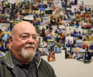 John Gallagher stands in front of a wall of photos of emergency medical service workers and volunteers at Central Lincoln County Ambulance Service in Damariscotta. After half a century with the service, Gallagher will retire in early 2022. (Emily Hayes photo)
