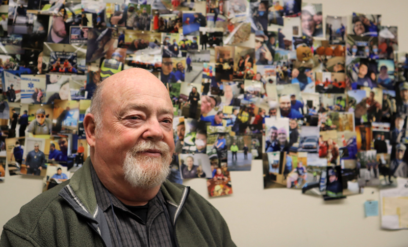 John Gallagher stands in front of a wall of photos of emergency medical service workers and volunteers at Central Lincoln County Ambulance Service in Damariscotta. After half a century with the service, Gallagher will retire in early 2022. (Emily Hayes photo)