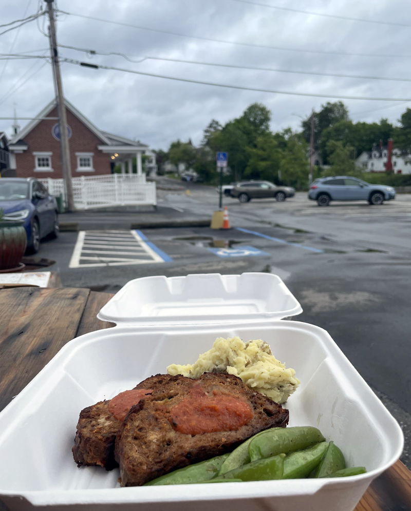 A view of a takeout order of Annettes meatloaf marinara from Savory Maine in Damariscotta, with the white building of the restaurant showing in the distant background, on Sunday, June 19. The dish was the first recipe Savory Maines owner and chef, Grace Goldberg, learned from her mother when she was young and it is still on the menu. (Evan Houk photo)