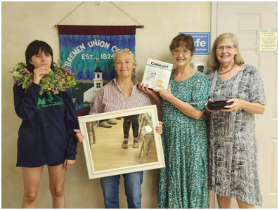 From left: Ireland Teele displays a wreath and samples one of the locally made pastries; Barbara Caggini displays a framed mirror, Joan Teele shows a kitchen appliance, and Janet Leonard holds the rest of the cookies. (Photo courtesy Bremen Union Church)