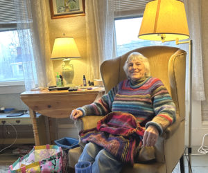 Patsy Dufresne, of Damariscotta, sits in her living room wearing one of her "traveling sweaters" with another in-progress sweater in her lap. Dufresne estimated that she has made 160 sweaters in the last 10 years, giving most away to friends and family. (Molly Rains photo)