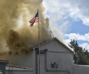 An American flag flutters above the warehouse that formerly housed William Smith Enterprises at 147 Somerville Road in Jefferson, while firefighting crews from at least 10 towns work below to quench the fire that consumed part of the building on the afternoon of Thursday, Aug. 22. (Molly Rains photo)