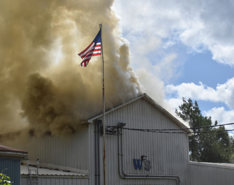 An American flag flutters above the warehouse that formerly housed William Smith Enterprises at 147 Somerville Road in Jefferson, while firefighting crews from at least 10 towns work below to quench the fire that consumed part of the building on the afternoon of Thursday, Aug. 22. (Molly Rains photo)