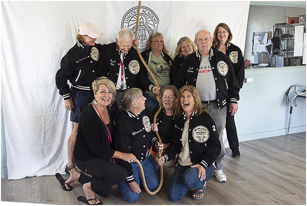 The 1982 state championship girls basketball team gather to ring the bell wearing their championship jackets. Front row, from left:  Cindy (Yeaton) Wade, Biz Williamson, Hannah (Russell) Laday, and Traci (Nelson) Halvorson. Back row, from left: Kim (McFarland) Bailey, coach Walt White, Britt Hatch, Lori (Belknap) McCullagh, coach Dan Pinkham, and Heather OBryan. (Photo courtesy Lincoln Academy)