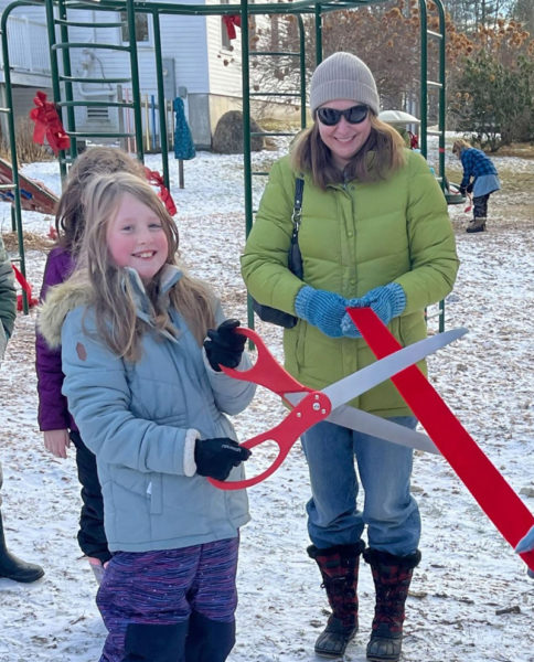 Damariscotta Montessori School student Maia Greenleaf takes a turn cutting the ribbon with the giant scissors during a small ribbon-cutting ceremony celebrating the addition of four new pieces of equipment the schools playground. The new additions are a new set of swings, a spinning structure, a large climbing structure with monkey bars, and a smaller rock wall structure. (Photo courtesy Damariscotta Montessori School)