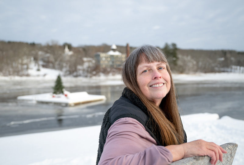 Rebecca Smith Waddell smiles from a stone bench at the snow-covered Pine Street Landing in Waldoboro on Saturday, Dec. 27. Behind her the towns floating Christmas tree is hemmed in by ice on the Medomak River. (Bisi Cameron Yee photo)