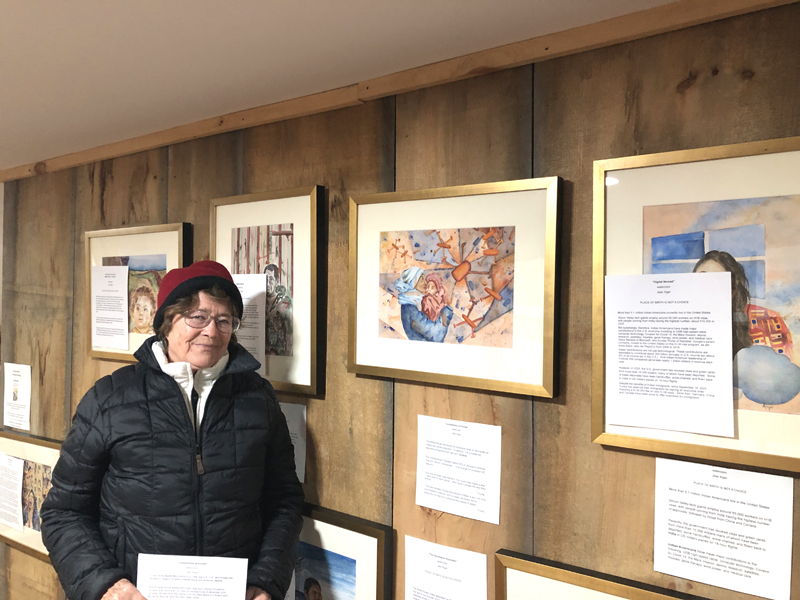 Waldoboro artist Jean Kigel poses in her Back Cove gallery next Constellation of Drones, one of 11 portraits she created to capture various current immigrant experiences in the United States and beyond in Displacement: Immigrant Portraits. The exhibition is on display through January at Skidompha Public Library in Damariscotta. (Sarah Masters photo)