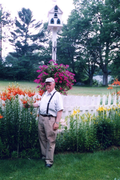 Calvin Dodge stands under a large hanging basket of petunias in full bloom. Above the petunias is an eight room church-style birdhouse he built in 1965, the year his son was born. The church birdhouse is now down in the backyard of his sons home in Manchester, Conn. In the summer of 2025 it had the nests of eight eastern bluebirds in it. (Photo courtesy Calvin Dodge collection)