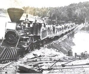 A gravel train at Muscongus Bay during construction of the Knox & Lincoln Railroad. The large crew of men on the flatbed cars were employed in hand-loading fill for the roadbed. This train was crossing a temporary spur track built on cobbled timbers across the waters of Muscongus Bay. Note the wood burning steam engine. (Courtesy photo)