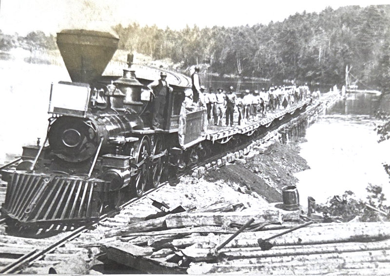A gravel train at Muscongus Bay during construction of the Knox & Lincoln Railroad. The large crew of men on the flatbed cars were employed in hand-loading fill for the roadbed. This train was crossing a temporary spur track built on cobbled timbers across the waters of Muscongus Bay. Note the wood burning steam engine. (Courtesy photo)