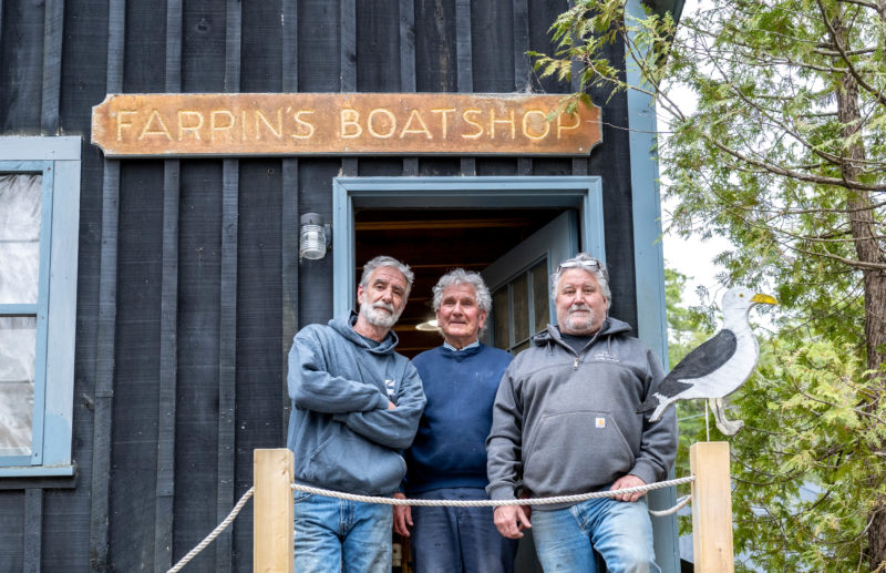 Brian Farrin (left), Bruce Farrin (center), and Bruce Farrin Jr. stand at the entrance to Farrins Boatshop in South Bristol on April 15. Bruce Farrin built the boatshop at this location in 1978 with the help of his wife and his sons grew up working in the yard. (Bisi Cameron Yee photo)